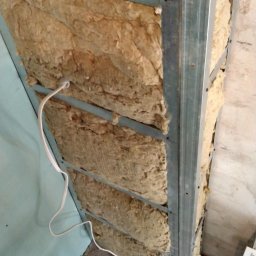 Kipish-Master Mykola Choban - Montaż izolacji z wełny mineralnej w metalowym stelażu, widoczne przewody elektryczne i fragment ściany w kolorze jasnoniebieskim.