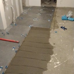 Kipish-Master Mykola Choban - Proces układania szarych płytek podłogowych, z widoczną warstwą kleju na podłodze i niebieskimi krzyżykami dystansowymi, w tle widoczna osoba w krótkich spodenkach.