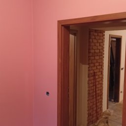 Kipish-Master Mykola Choban - Świeżo pomalowana na różowo ściana z widocznymi otworami na gniazdka elektryczne, drewniana futryna drzwi i ceglana ściana w tle, z rozłożonym kablem zasilającym i butelką wody mineralnej w rogu.