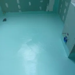 Świeżo nałożona, turkusowa hydroizolacja w łazience, widoczne ślady pędzla na podłodze, przygotowane ściany z płyt gipsowo-kartonowych, niebieska rura kanalizacyjna wystająca ze ściany.