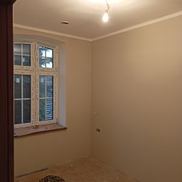 Kipish-Master Mykola Choban - Pokój po malowaniu ścian na beżowo, widoczna puszka farby Greinplast, okno z białą ramą, zakryte podłogi, surowa żarówka.