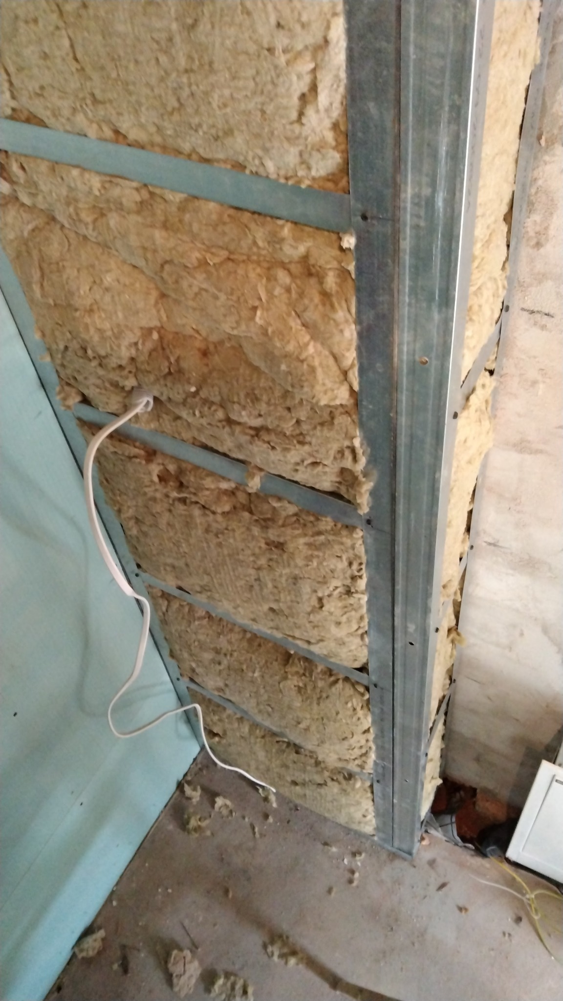 Montaż izolacji z wełny mineralnej w metalowym stelażu, widoczne przewody elektryczne i fragment ściany w kolorze jasnoniebieskim.