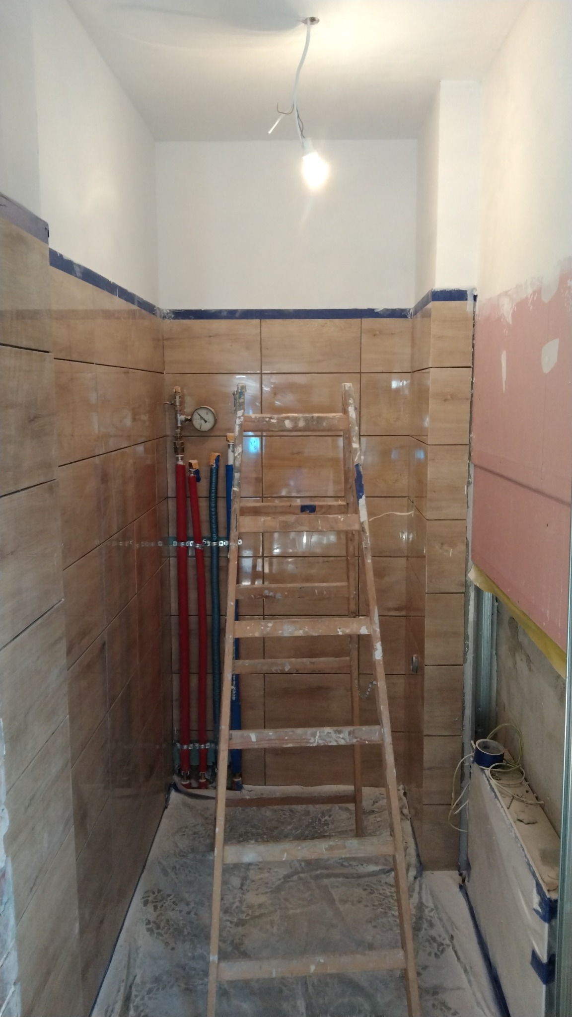 Wnętrze łazienki w trakcie wykańczania: położone brązowe płytki imitujące drewno, widoczne rury instalacji wodnej z manometrem, drewniana drabina malarska, zabezpieczona podłoga i fragment ściany...
