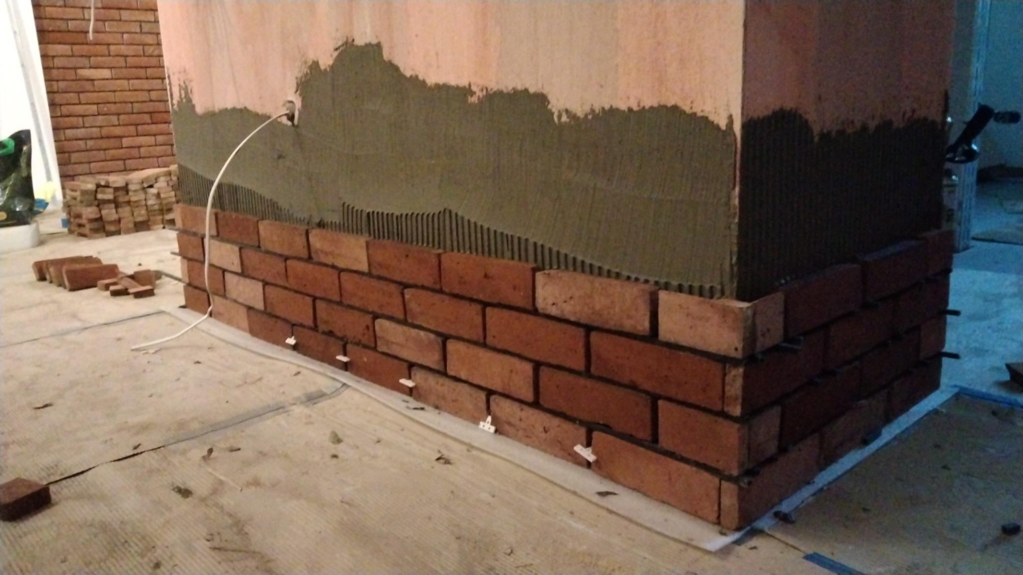 Fragment ściany z częściowo ułożoną cegłą klinkierową, widoczny klej na ścianie i plastikowe krzyżyki dystansowe, w tle stos cegieł.