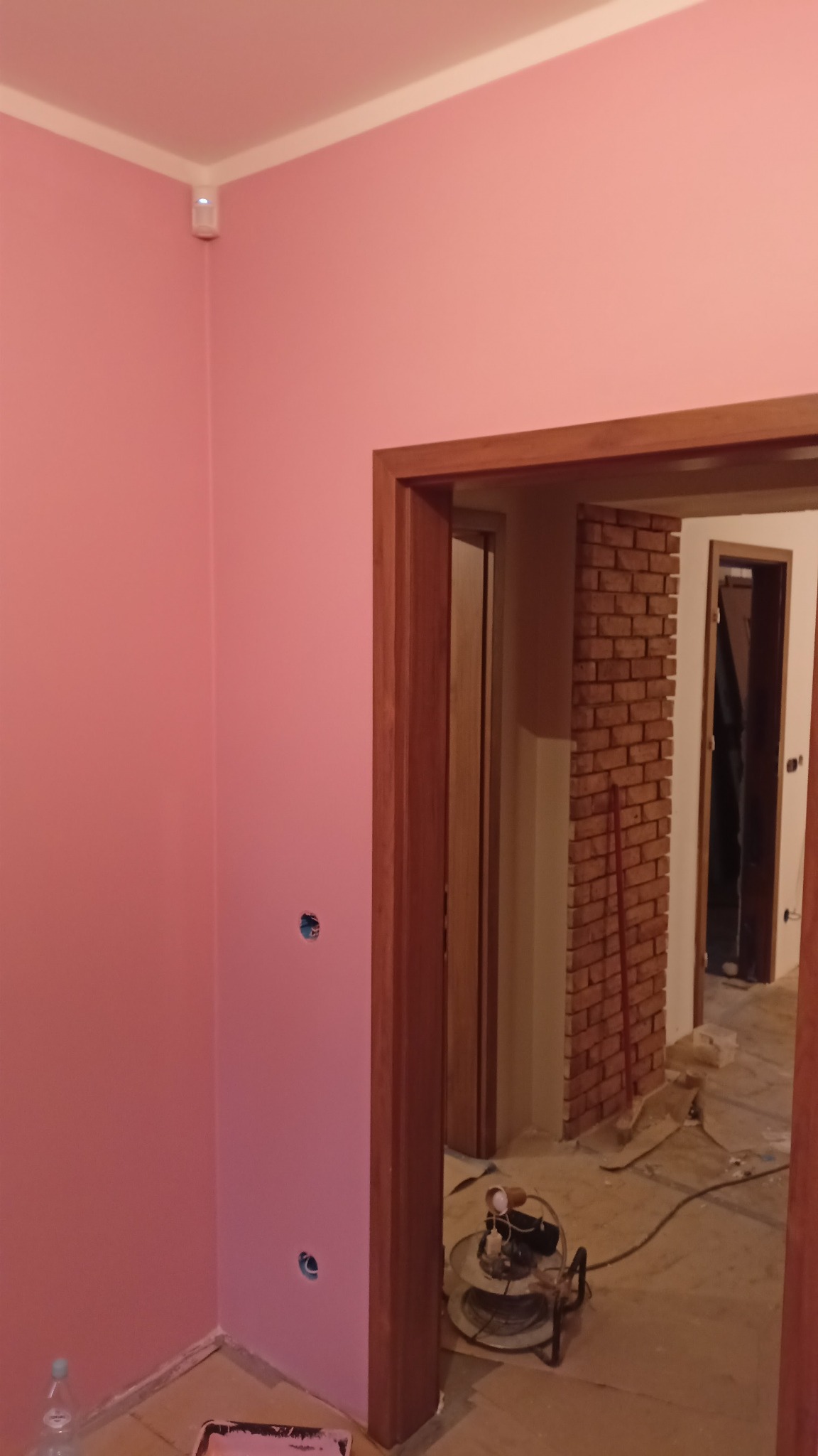 Świeżo pomalowana na różowo ściana z widocznymi otworami na gniazdka elektryczne, drewniana futryna drzwi i ceglana ściana w tle, z rozłożonym kablem zasilającym i butelką wody mineralnej w rogu.