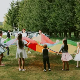 Grupa dzieci i dorosłych bawi się na trawie z kolorową chustą animacyjną, w tle widoczne drzewa i zaparkowane samochody.