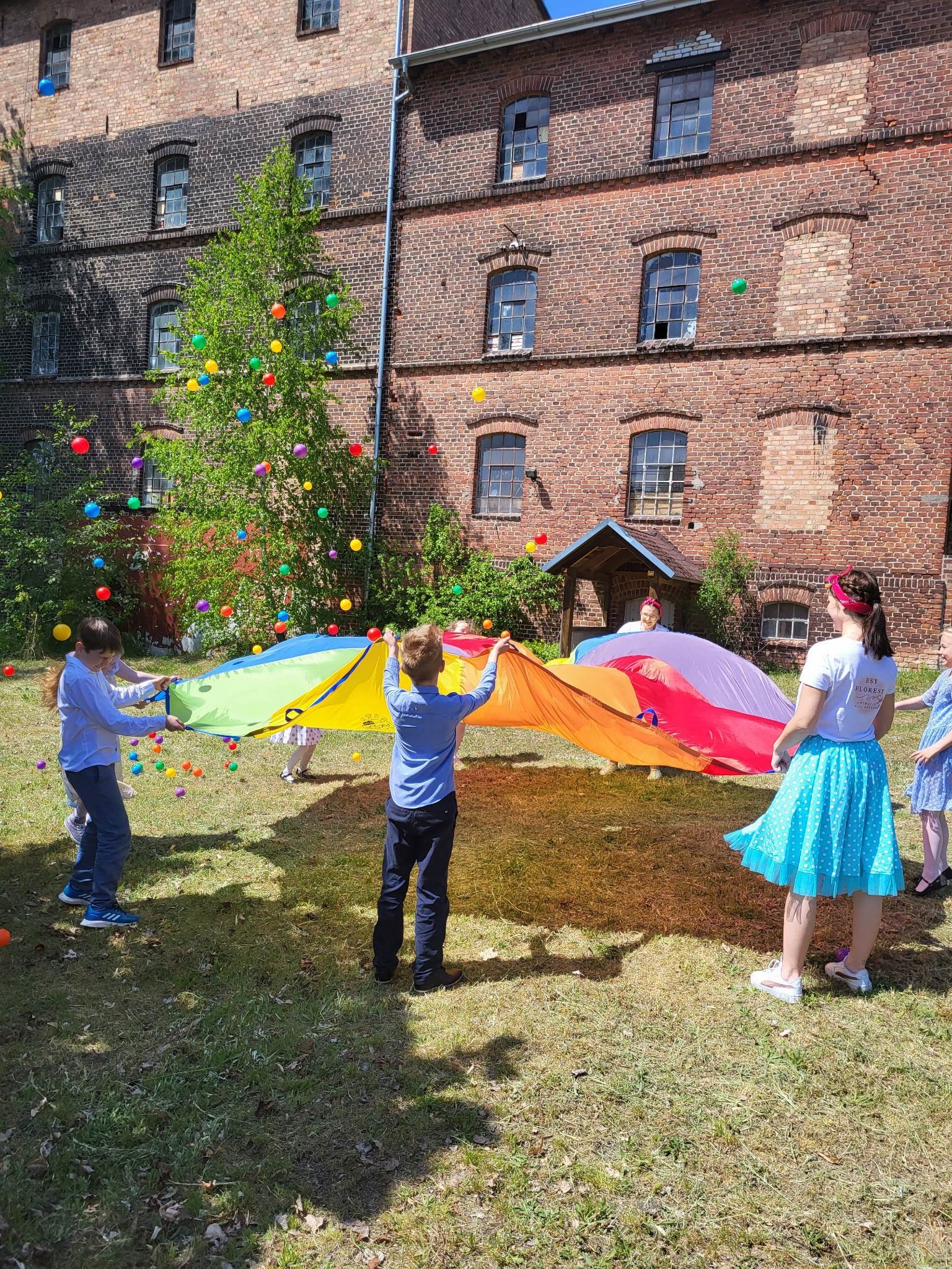 Grupa dzieci i dorosłych bawi się kolorową chustą animacyjną i unoszącymi się w powietrzu piłeczkami na tle ceglanego budynku.