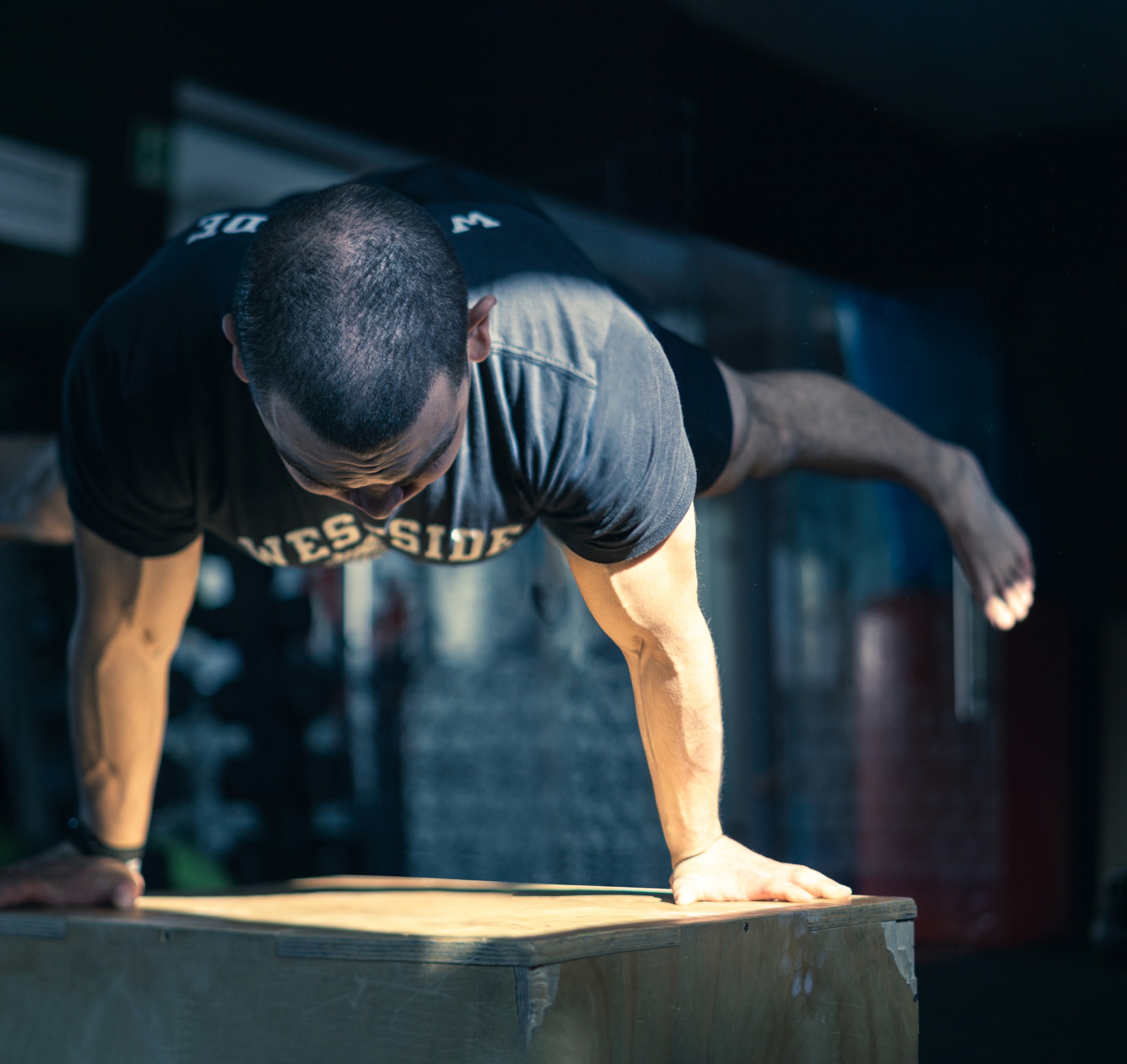Mężczyzna w koszulce z napisem 'WEST SIDE' wykonuje ćwiczenie stania na rękach na drewnianej skrzyni w ciemnym pomieszczeniu, widoczne napięte mięśnie ramion i brak obuwia.