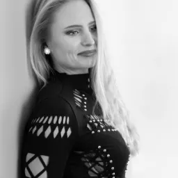 Portret kobiety opierającej się o ścianę, w czarno-białej tonacji, ubrana w bluzkę z wycięciami i srebrnymi nitami, z kolczykami w kształcie kwiatów, delikatny uśmiech.