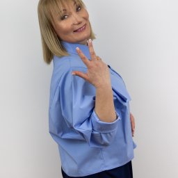 MARTA KOTECKA - Portret uśmiechniętej kobiety w błękitnej koszuli, pozującej na białym tle w Posłusze, z uniesioną dłonią pokazującą trzy palce i subtelnym makijażem.