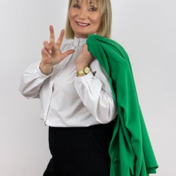 MARTA KOTECKA - Uśmiechnięta blond kobieta w białej koszuli i czarnych spodniach, z zieloną marynarką na ramieniu, pokazuje trzy palce na białym tle.
