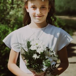 MARTA KOTECKA - Uśmiechnięta dziewczynka w białej sukience i wianku z drobnych kwiatów trzyma bukiet białych kwiatów na tle zieleni.