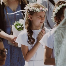 MARTA KOTECKA - Dziewczynka w białej sukience komunijnej z wiankiem na głowie, z złożonymi dłońmi, uśmiecha się w tłumie, w tle widoczni rodzice i goście.