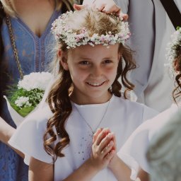 MARTA KOTECKA - Uśmiechnięta dziewczynka w białej sukience komunijnej i wianku z białych i różowych kwiatów, z dłońmi złożonymi do modlitwy, otoczona przez rodzinę.