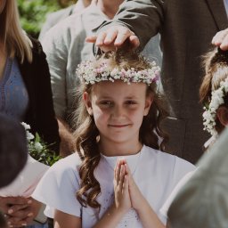 MARTA KOTECKA - Dziewczynka w białej sukience komunijnej i wianku z białych i różowych kwiatów, z rękami złożonymi do modlitwy, otoczona przez rodzinę podczas uroczystości.