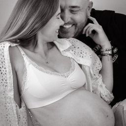 MARTA KOTECKA - Czarno-białe zdjęcie uśmiechniętej kobiety w bieliźnie i ażurowej koszuli, z widocznym ciążowym brzuchem, wtulonej w uśmiechniętego mężczyznę z brodą, oboje patrzą na siebie, w tle logo fotografa.