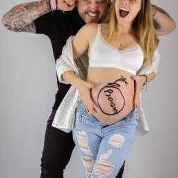 MARTA KOTECKA - Uśmiechnięta przyszła mama z napisem 'Boom' na brzuchu, trzymająca ręce na brzuchu, obok stoi tata z tatuażami na rękach, oboje w radosnych pozach na jasnym tle.