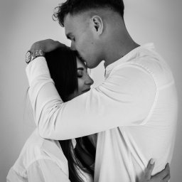 MARTA KOTECKA - Czarno-białe zdjęcie obejmującej się pary. Mężczyzna w jasnej koszuli całuje kobietę w głowę, ona ma zamknięte oczy. Na ręce mężczyzny widoczny zegarek.