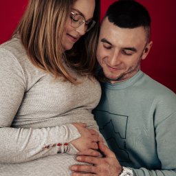 MARTA KOTECKA - Przyszli rodzice obejmujący ciążowy brzuszek na czerwonym tle, widoczny zegarek G-Shock na nadgarstku mężczyzny.