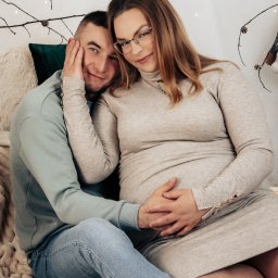 MARTA KOTECKA - Uśmiechnięta para w ciepłych ubraniach siedzi na puszystym kocu, mężczyzna trzyma dłoń na brzuchu kobiety w ciąży, w tle gałązki z lampkami i logo fotografa.