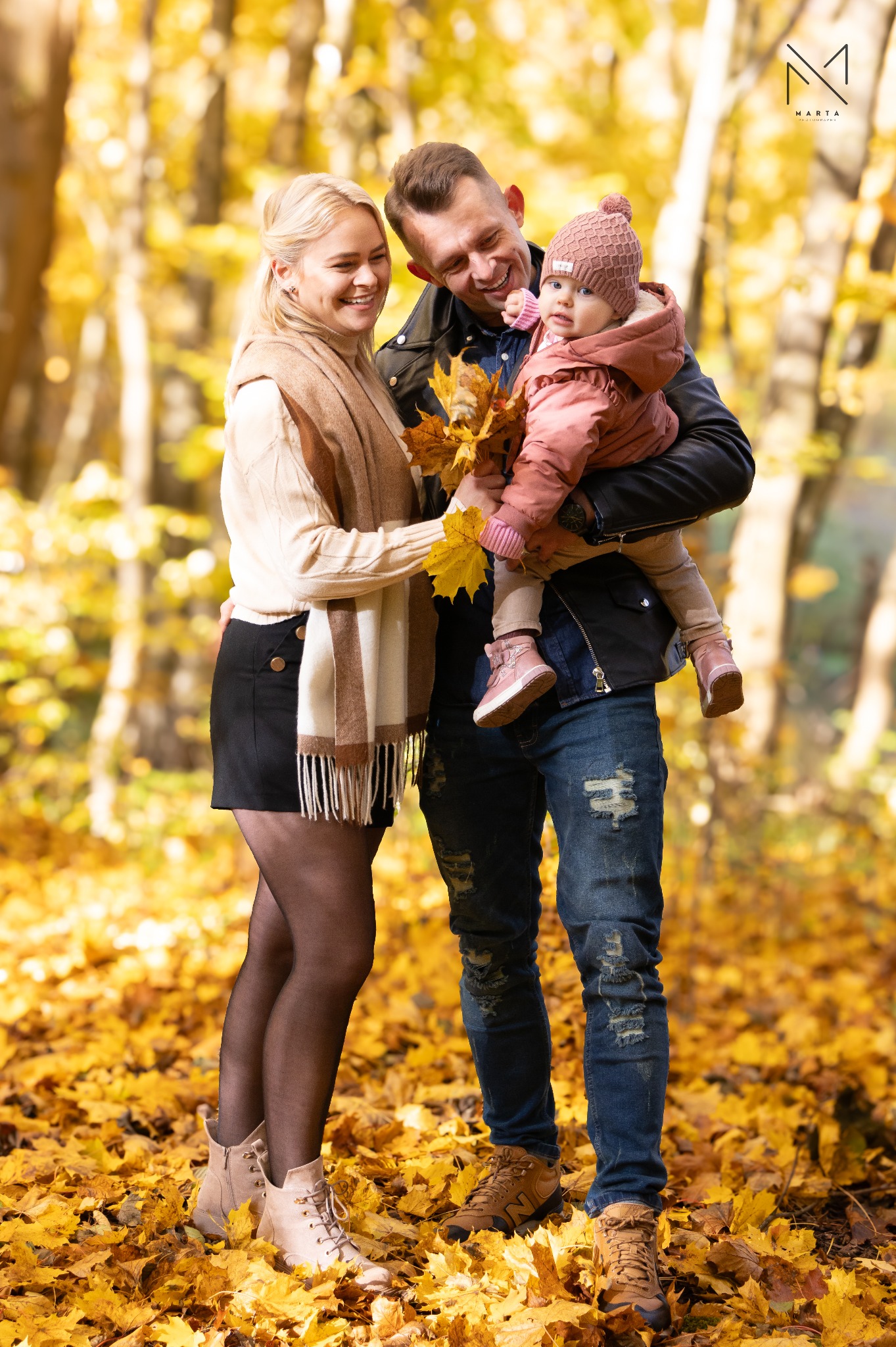 Uśmiechnięta rodzina z małym dzieckiem w jesiennym parku, kobieta trzyma garść żółtych liści, ciepłe światło słoneczne.