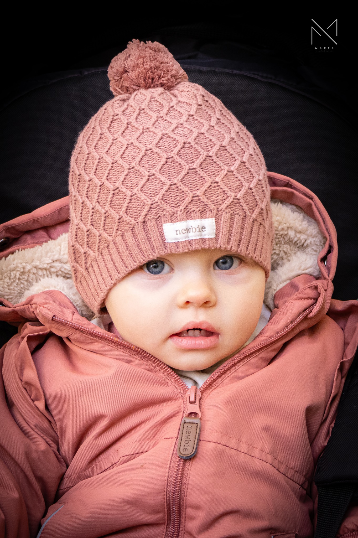 Portret niemowlęcia w różowej, pikowanej kurtce z kapturem i dzierganej czapce z pomponem, z niebieskimi oczami i lekko otwartymi ustami.