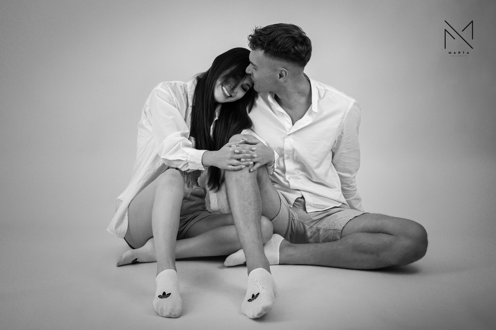 Czarno-białe zdjęcie uśmiechniętej kobiety opierającej głowę na ramieniu mężczyzny, oboje siedzą na podłodze w białych koszulach i krótkich spodenkach, ona w skarpetkach Adidas, w tle logo fotografa.