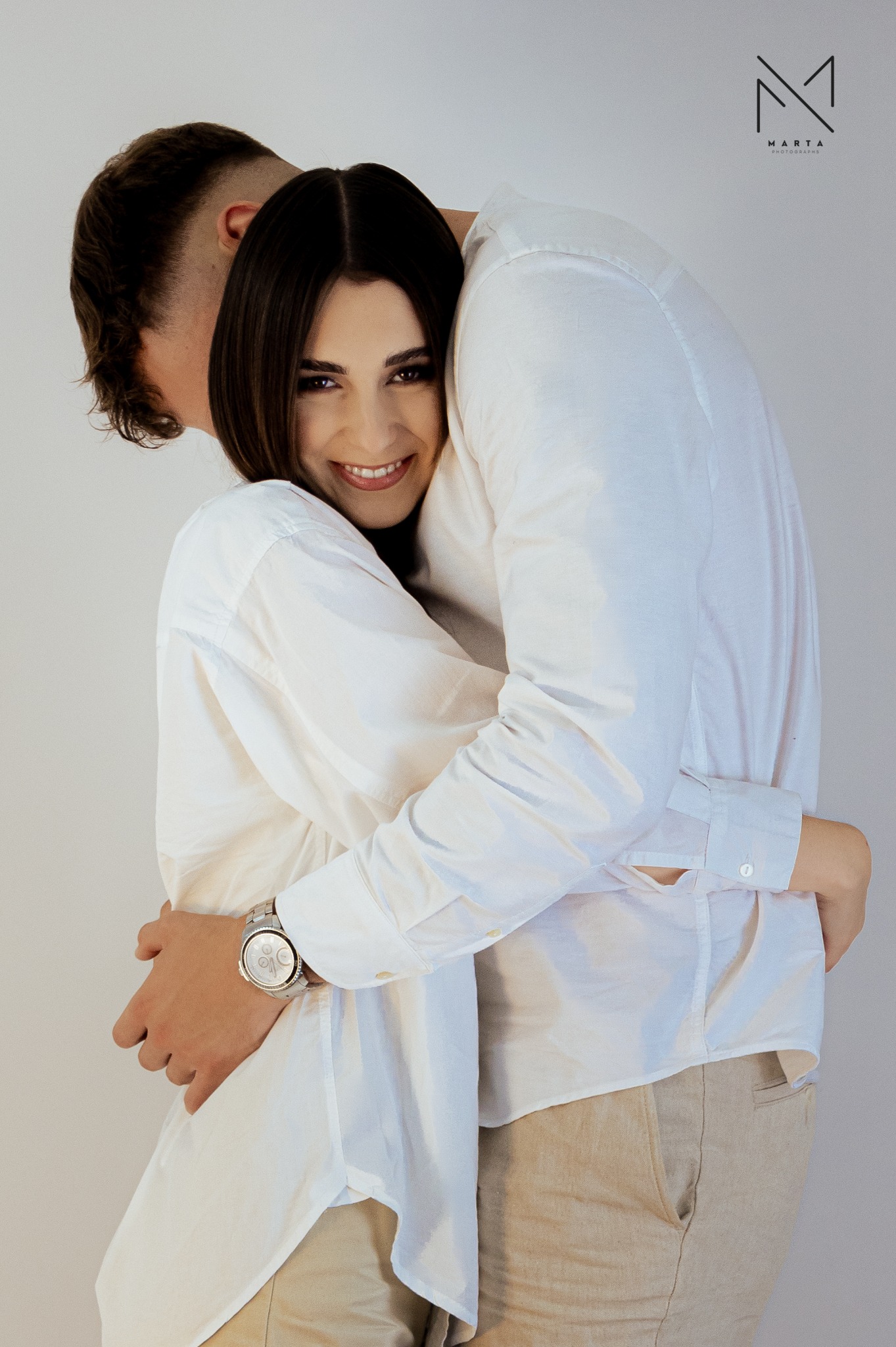 Uśmiechnięta kobieta w białej koszuli obejmowana przez mężczyznę w Posłusze, widoczny zegarek na ręce mężczyzny i logo fotografa w prawym górnym rogu.