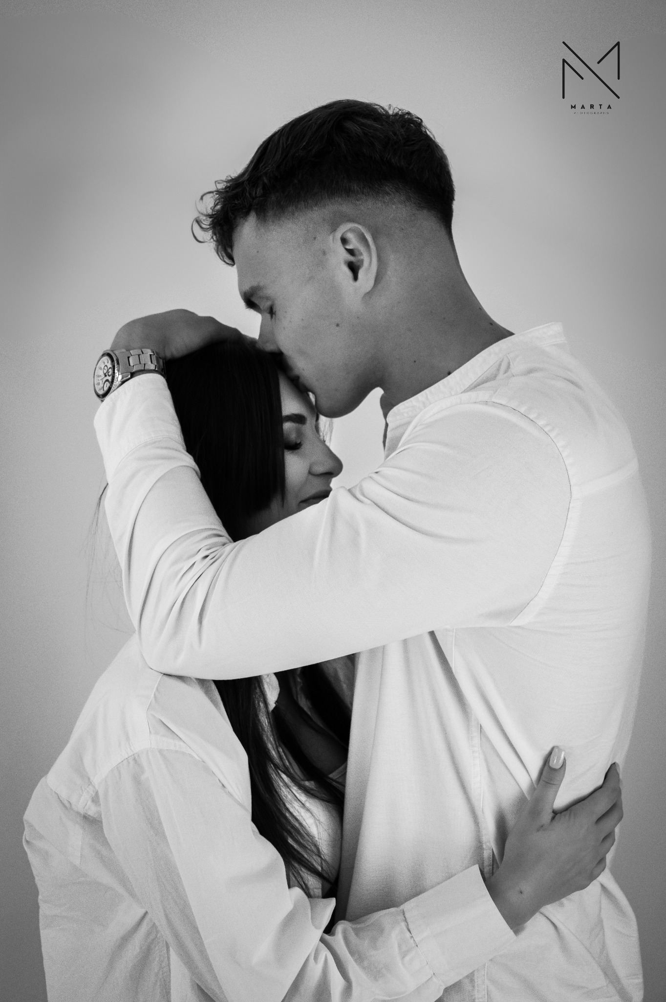 Czarno-białe zdjęcie obejmującej się pary. Mężczyzna w jasnej koszuli całuje kobietę w głowę, ona ma zamknięte oczy. Na ręce mężczyzny widoczny zegarek.