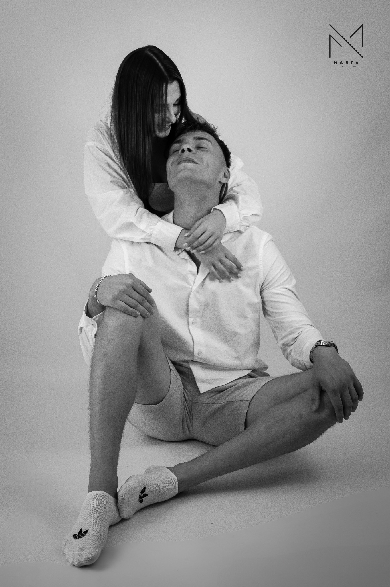 Czarno-białe zdjęcie pary: kobieta obejmuje siedzącego mężczyznę, oboje w jasnych koszulach, on w szortach i skarpetkach z logo Adidas, ona z długimi, prostymi włosami, w tle logo fotografa.