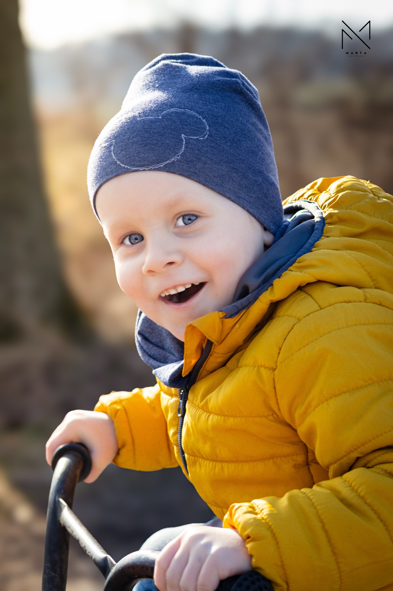 Uśmiechnięty chłopiec w żółtej kurtce i niebieskiej czapce z motywem Myszki Miki, trzymający się metalowej konstrukcji na tle rozmytej zieleni.