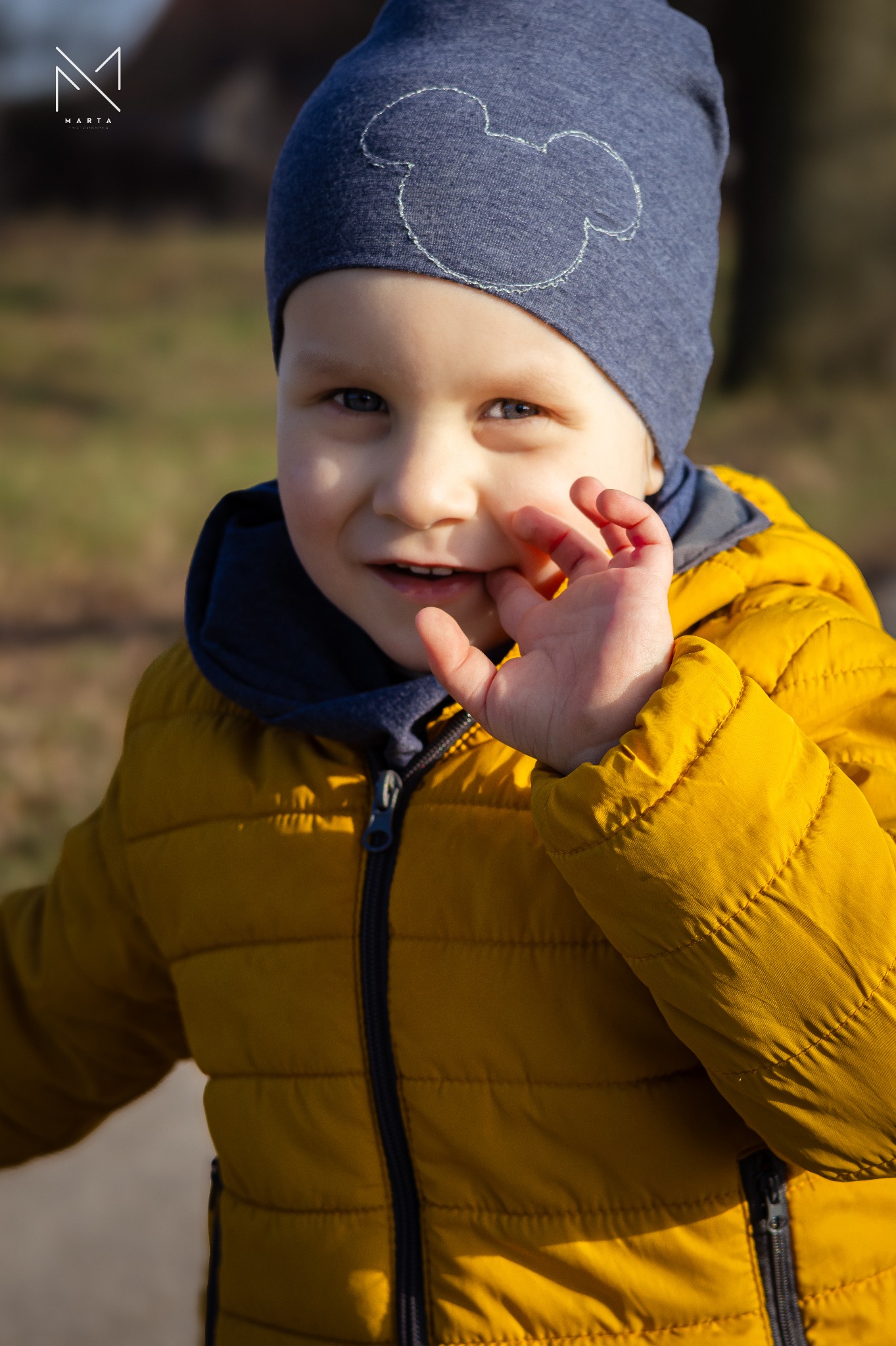 Uśmiechnięty chłopiec w żółtej kurtce i niebieskiej czapce z motywem Myszki Miki, trzymający palec w ustach na tle drzew.