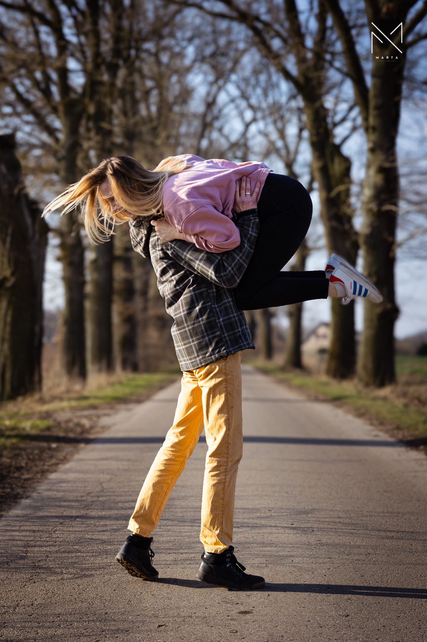 Mężczyzna w żółtych spodniach i kraciastej koszuli trzyma na rękach blondynkę w różowej bluzie na tle alei drzew.