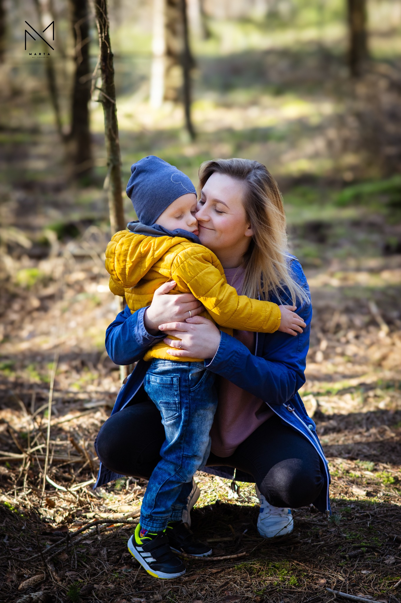 Matka w niebieskiej kurtce przytula synka w żółtej kurtce i niebieskiej czapce w lesie, delikatne światło słoneczne.