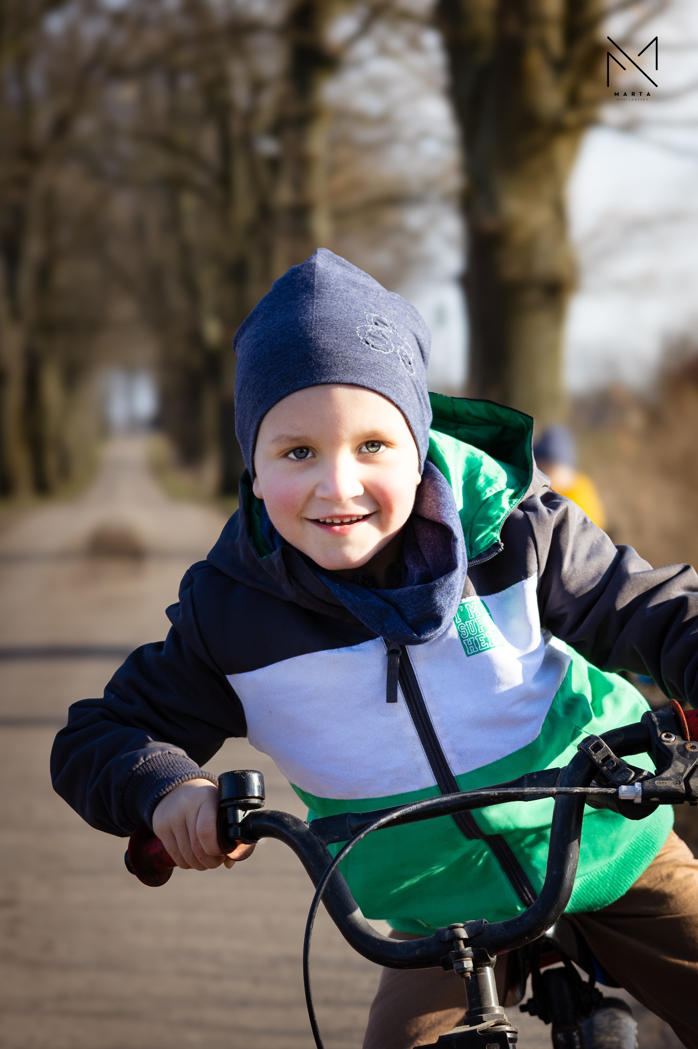 Uśmiechnięty chłopiec w czapce i kolorowej kurtce jedzie na rowerze po alei drzew, w tle rozmyty krajobraz, logo fotografa w prawym górnym rogu.