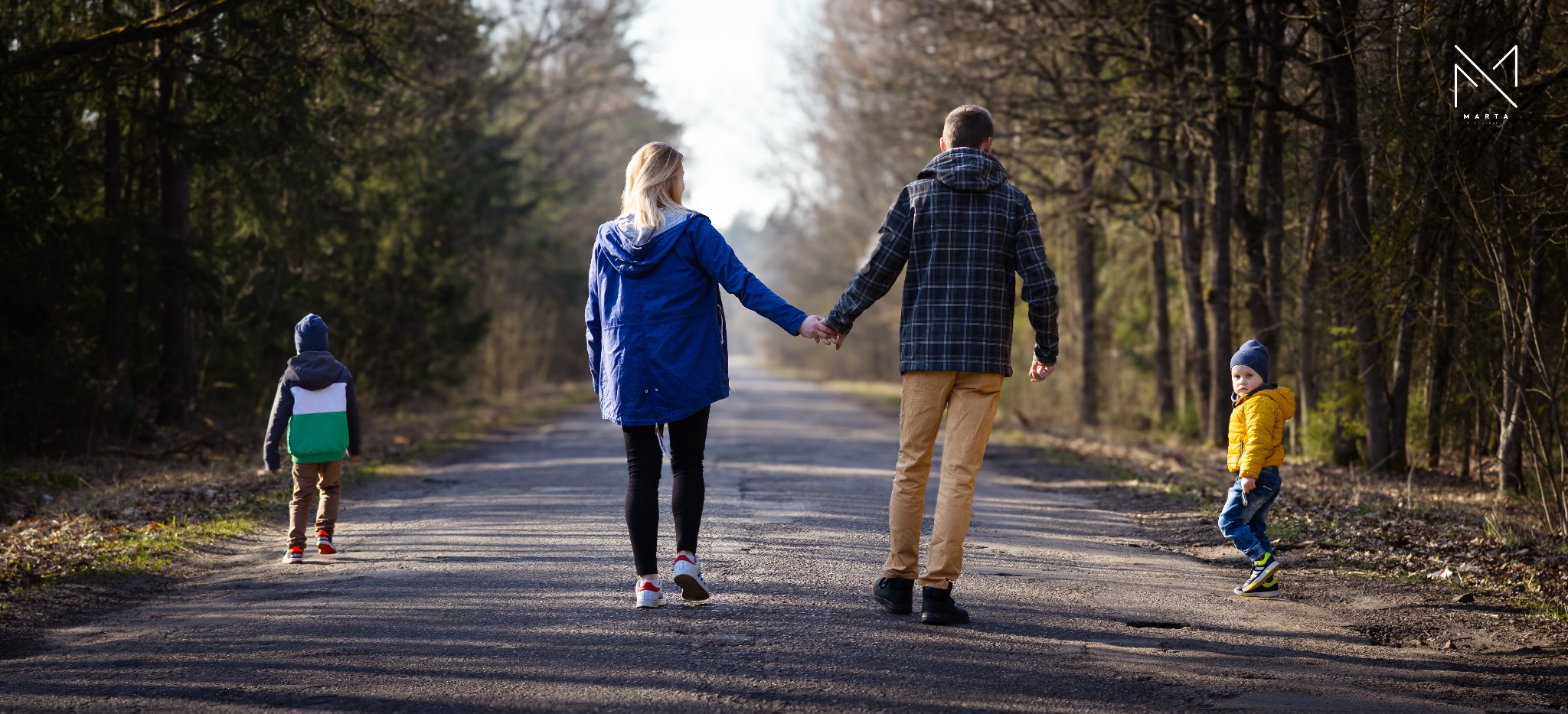 Rodzina spacerująca leśną drogą: rodzice trzymają się za ręce, syn z przodu w żółtej kurtce, drugi syn z tyłu w zielono-białej kurtce, widok z tyłu, w tle drzewa.