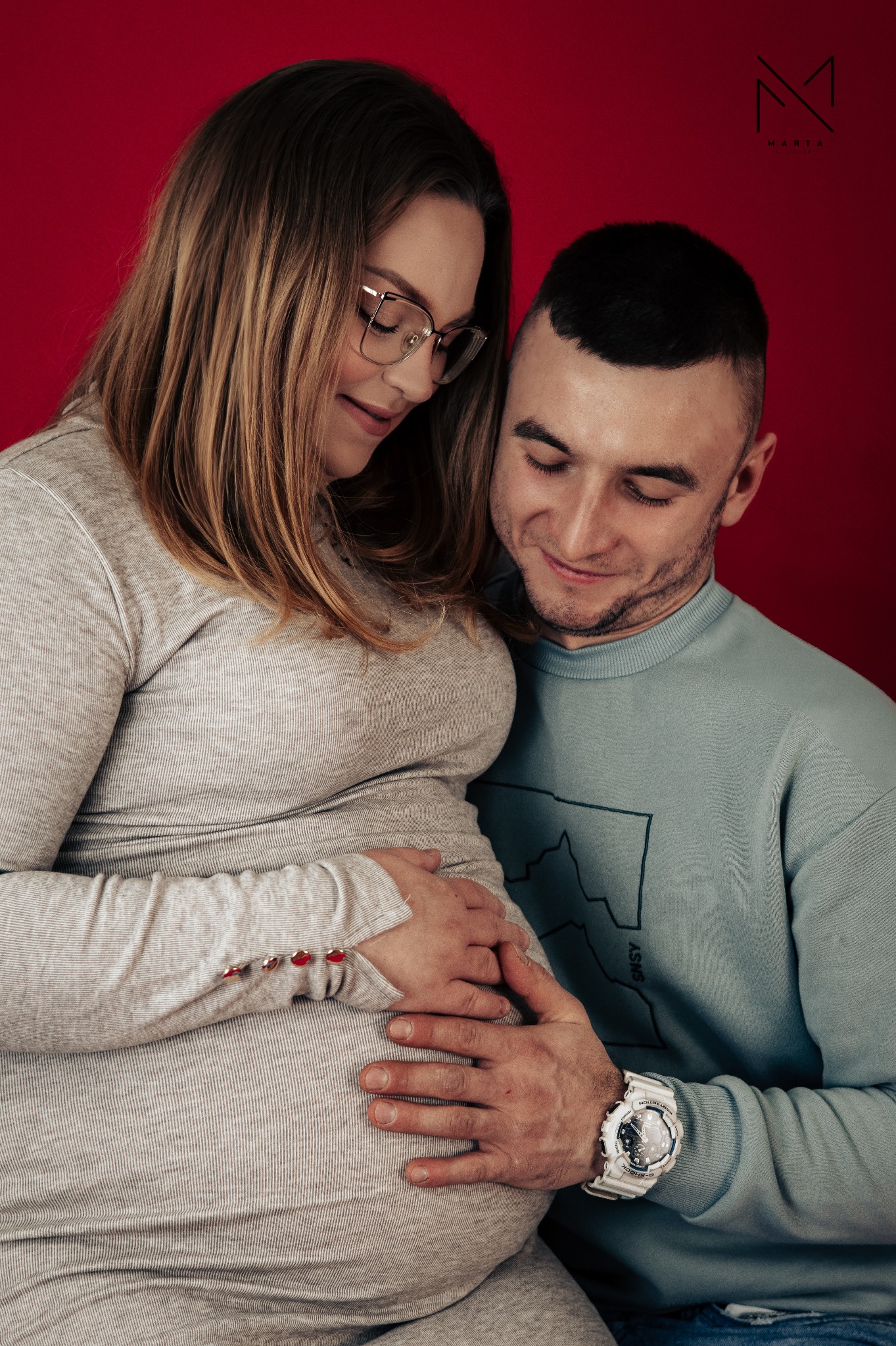 Przyszli rodzice obejmujący ciążowy brzuszek na czerwonym tle, widoczny zegarek G-Shock na nadgarstku mężczyzny.