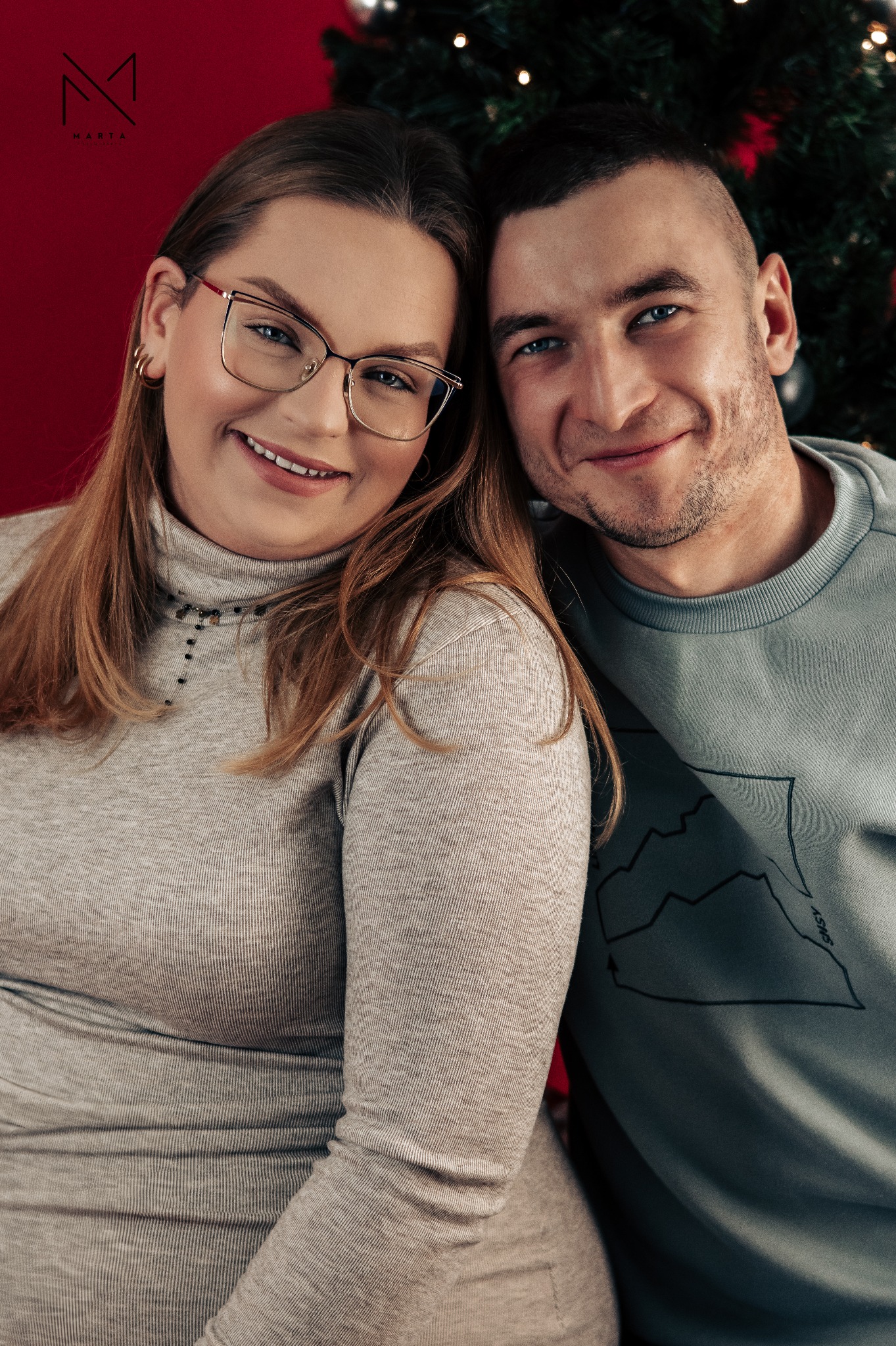 Portret uśmiechniętej pary, kobieta w okularach i golfie z ozdobnymi kamieniami, mężczyzna w bluzie z nadrukiem gór, na tle czerwonej ściany i choinki z lampkami.