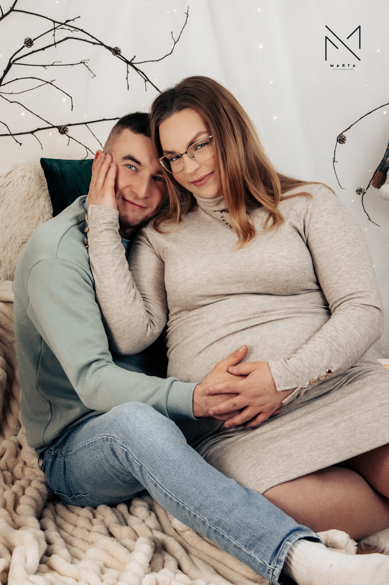 Uśmiechnięta para w ciepłych ubraniach siedzi na puszystym kocu, mężczyzna trzyma dłoń na brzuchu kobiety w ciąży, w tle gałązki z lampkami i logo fotografa.