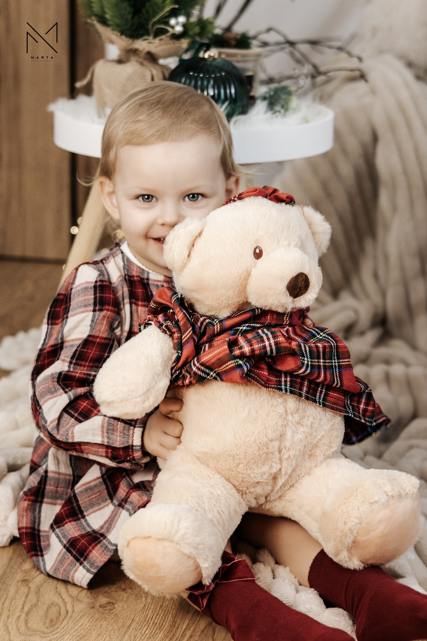 Uśmiechnięta dziewczynka w kraciastej sukience przytula pluszowego misia w kraciastej szarfie, siedząc na podłodze z drewna, w tle dekoracje świąteczne i puszysty koc.