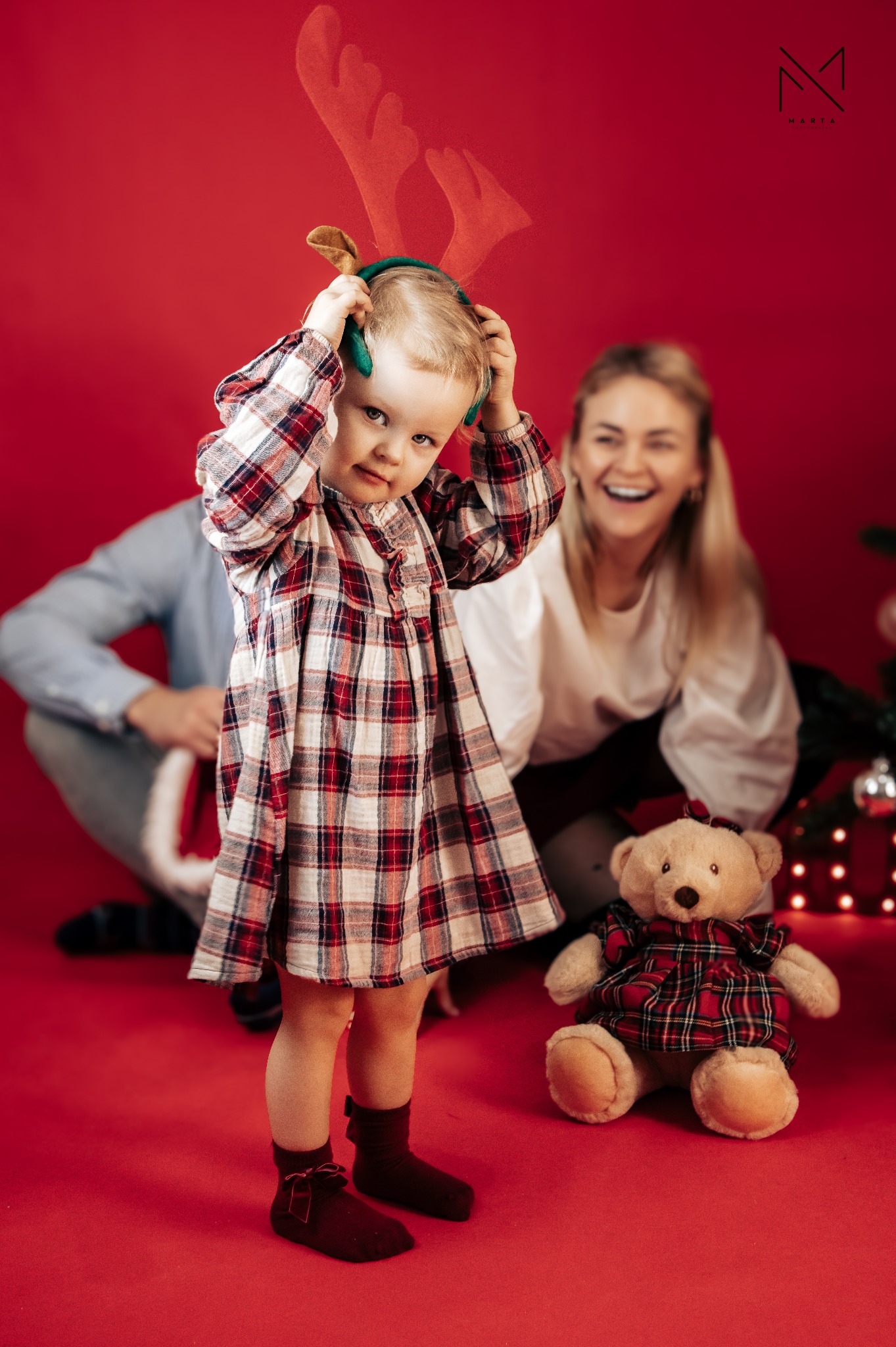 Dziewczynka w sukience w kratę i opasce z rogami renifera, poprawia ją na głowie. W tle uśmiechnięta kobieta i fragment choinki z prezentami na czerwonym tle.