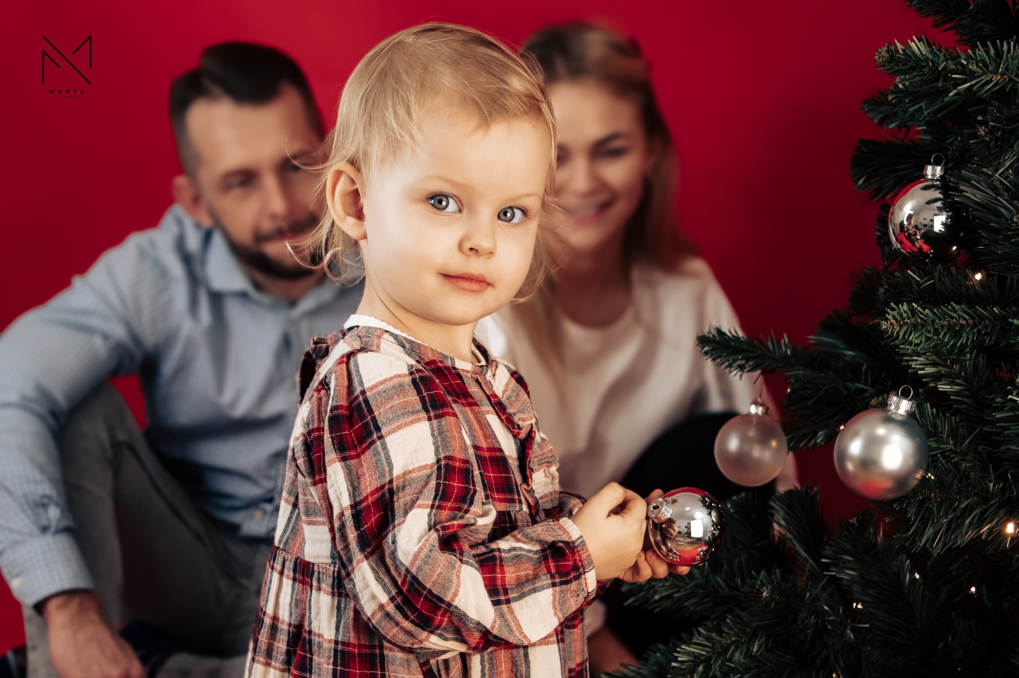 Dziewczynka w sukience w kratę trzyma srebrną bombkę na tle choinki i uśmiechniętej rodziny na czerwonym tle.