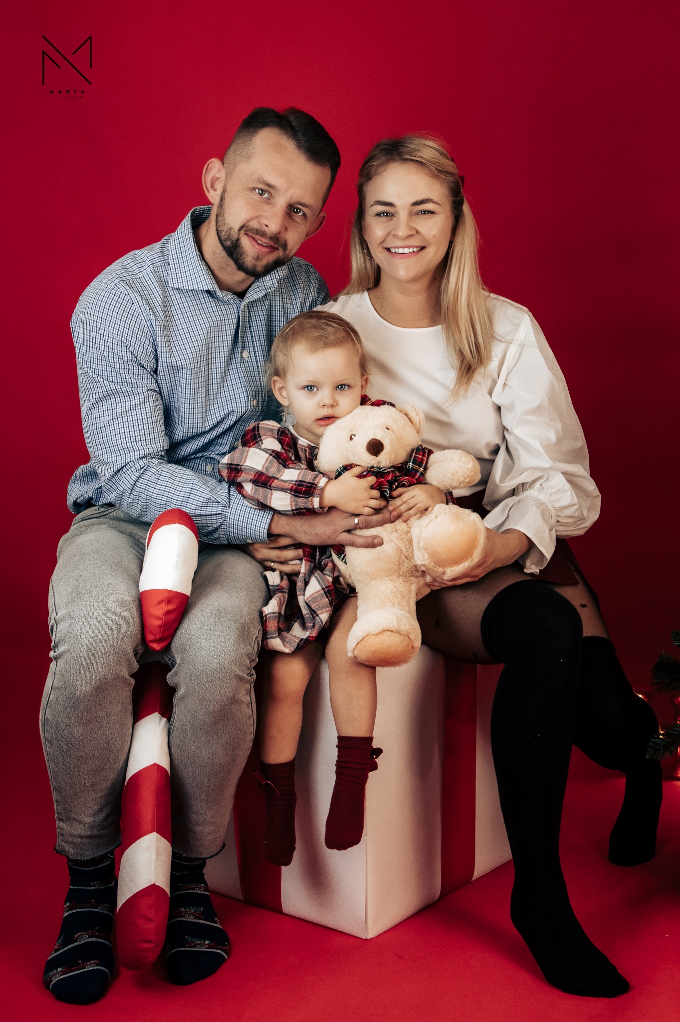 Rodzinne zdjęcie na czerwonym tle: uśmiechnięci rodzice trzymają dziecko z pluszowym misiem w świątecznej scenerii z rekwizytami.
