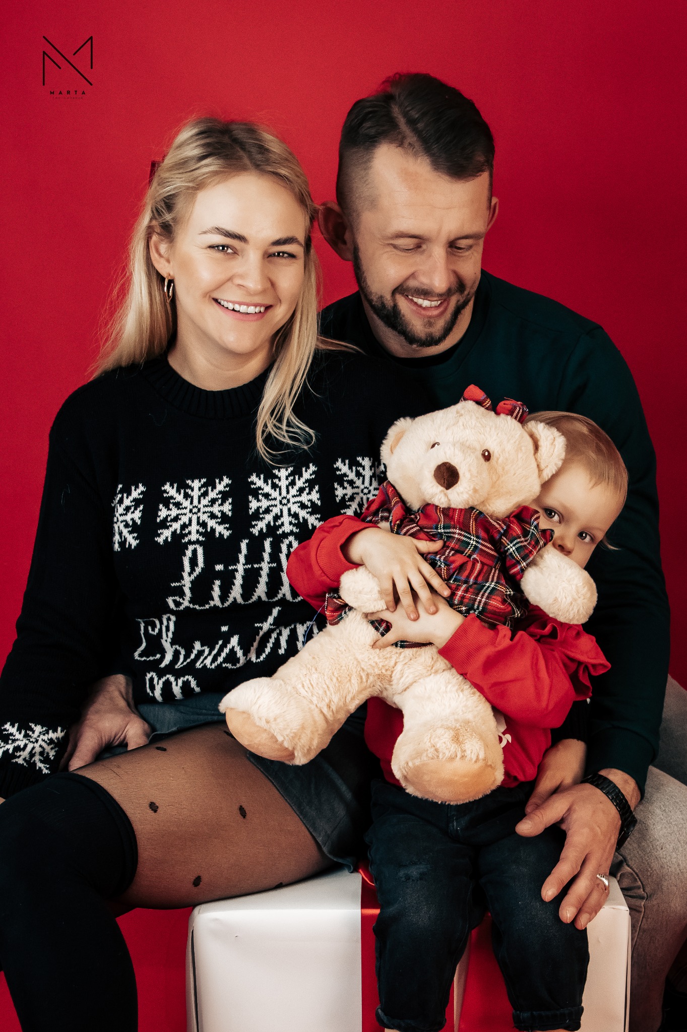 Uśmiechnięta rodzina z małym dzieckiem i pluszowym misiem w kratkę na czerwonym tle, sesja w świątecznym klimacie.