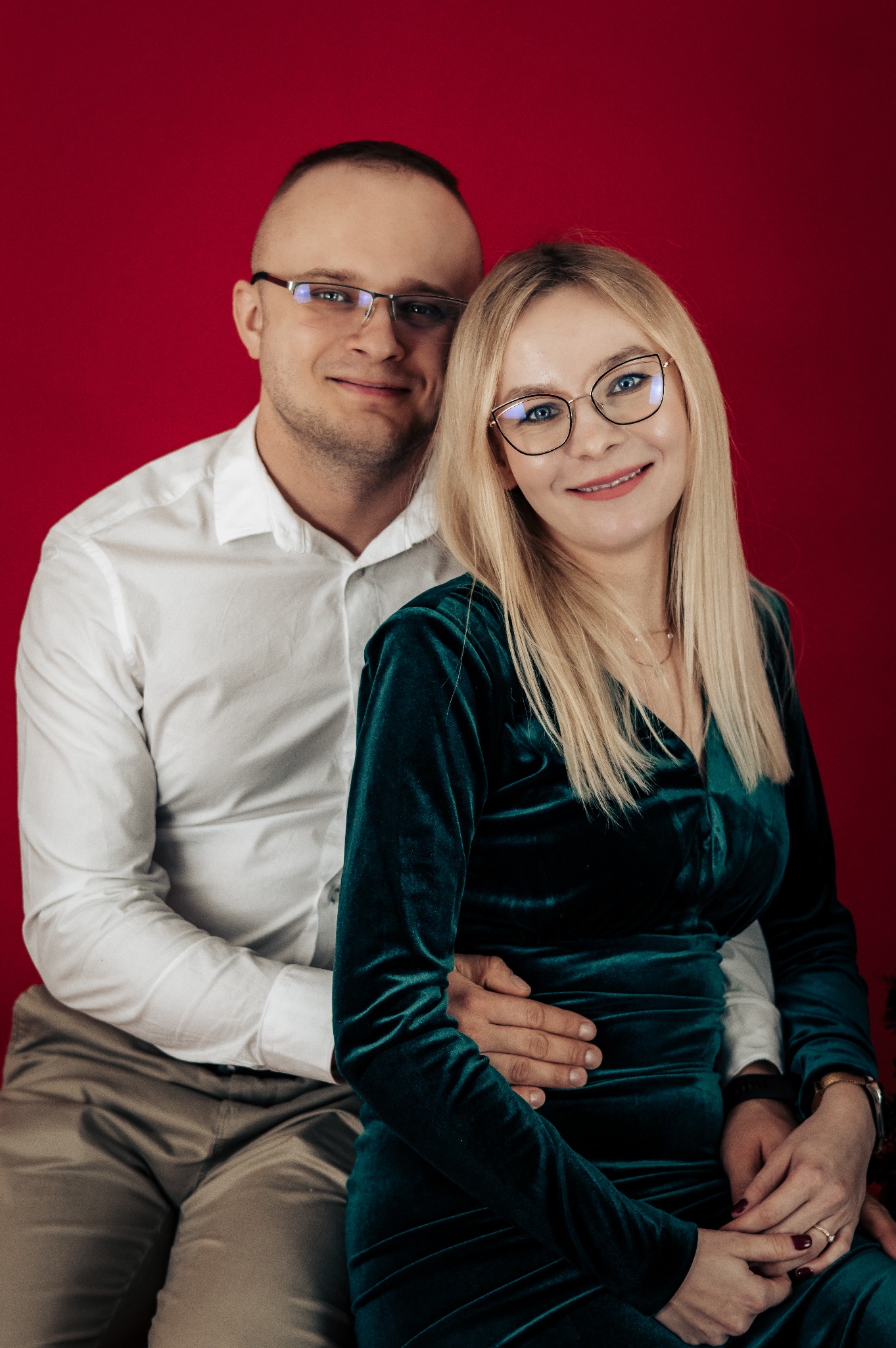Portret uśmiechniętej pary w okularach na czerwonym tle: mężczyzna w białej koszuli obejmuje kobietę w szmaragdowej, aksamitnej sukience.