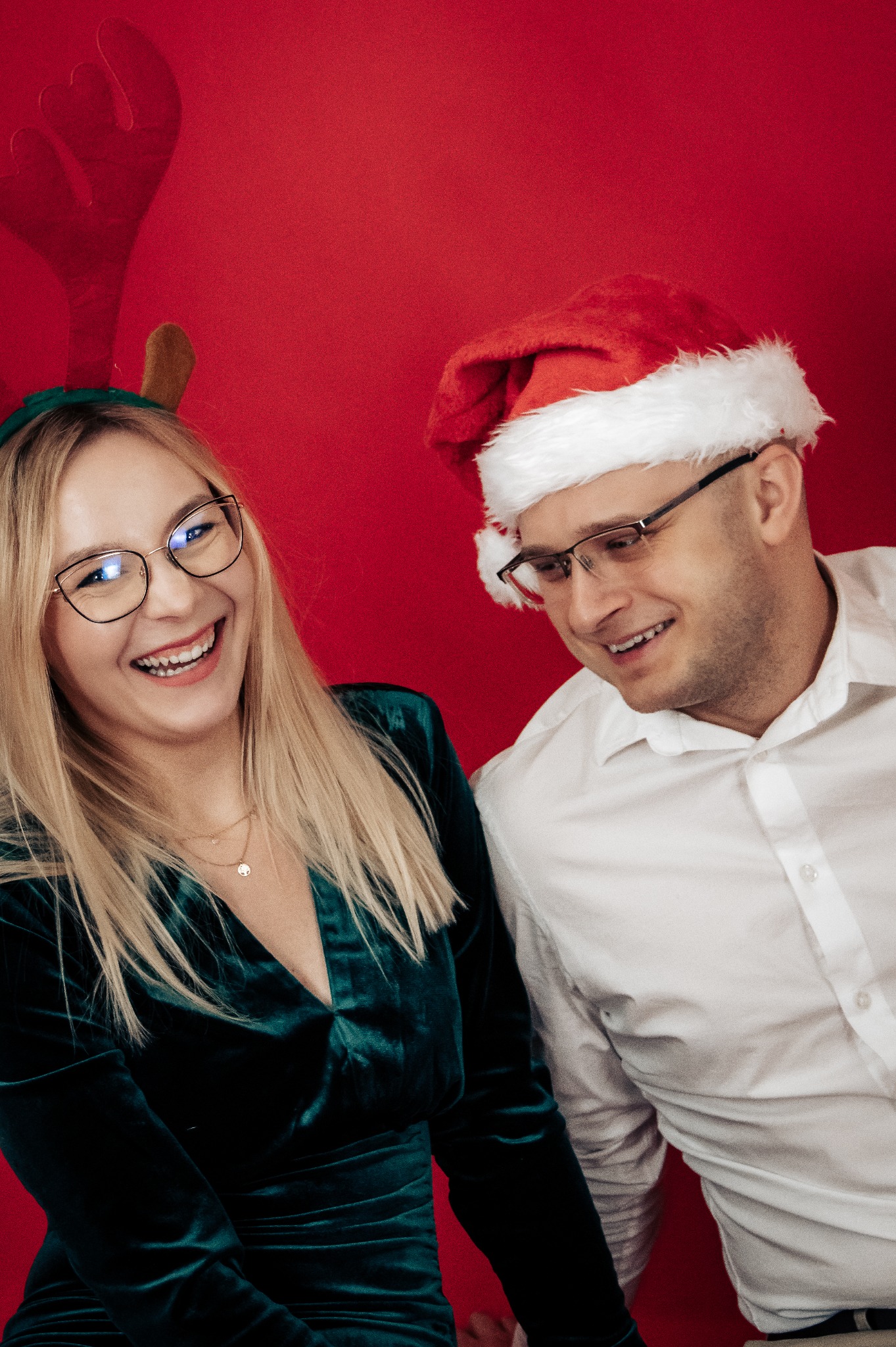 Uśmiechnięta para w świątecznych strojach na czerwonym tle: blondynka w zielonej sukience z rogami renifera i mężczyzna w białej koszuli i czapce Mikołaja, oboje w okularach.