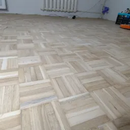 Ułożony parkiet w jodełkę francuską, widoczna maszyna do cyklinowania w tle oraz przedłużacz z białym gniazdkiem.