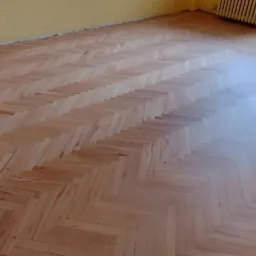 Świeżo wycyklinowany parkiet ułożony w jodełkę w pokoju z żółtymi ścianami, widoczne ślady po szlifowaniu przy ścianach.