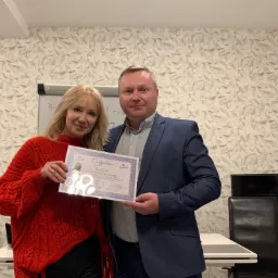 Kobieta w czerwonym swetrze i mężczyzna w niebieskiej marynarce trzymają certyfikat w pomieszczeniu z białą ścianą o geometrycznym wzorze.