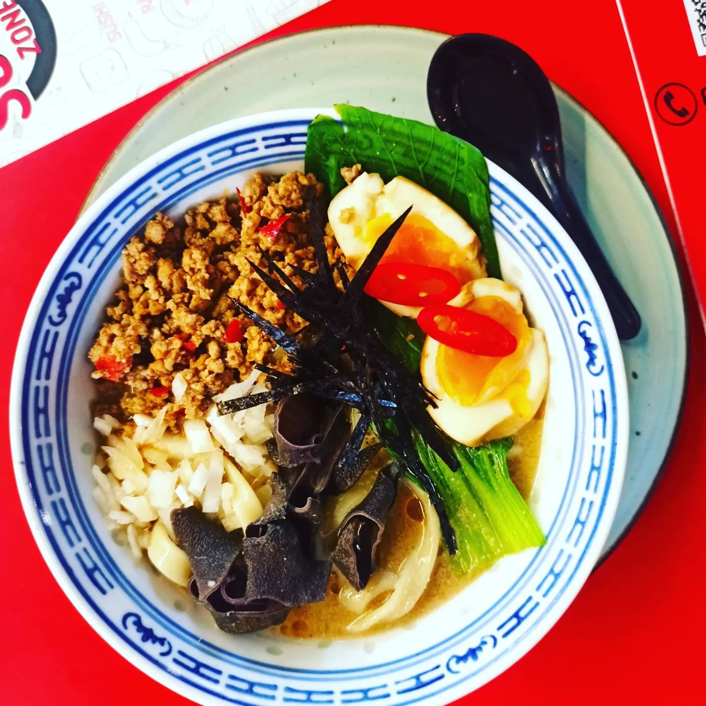 Ramen w ceramicznej misce z niebieskim wzorem, z jajkiem na miękko, grzybami mun, mielonym mięsem i papryczką chili, podany na talerzu na czerwonym tle.